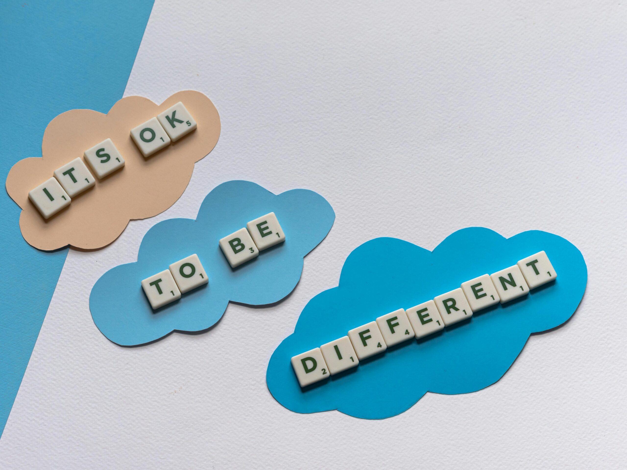 Scrabble tiles spell 'It's ok to be different' on colorful cloud cutouts, promoting diversity.
